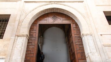 Tall pointed stone arch with partly open paneled wooden doors in a pale masonry wall.