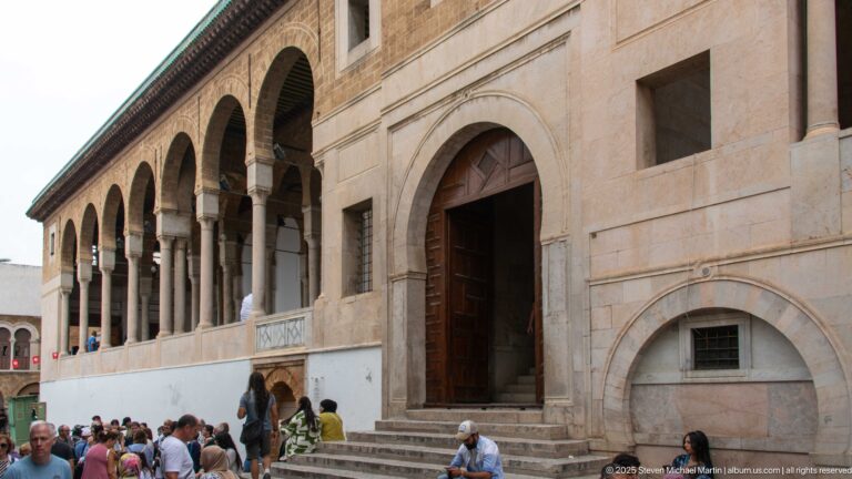 Visitors on stone steps at a tall arched entrance beneath an upper arcade on the Zitouna Grand Mosque exterior.