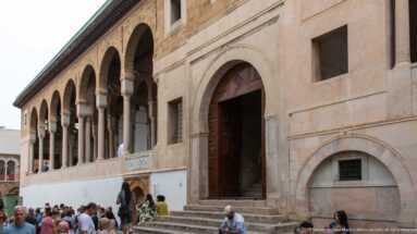 Visitors on stone steps at a tall arched entrance beneath an upper arcade on the Zitouna Grand Mosque exterior.
