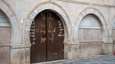 Dark double wooden door painted with gold and white Arabic calligraphy beneath a low stone arch.