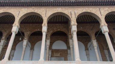 Two-tiered mosque courtyard arcade with pale columns, carved capitals, warm stone arches, and dark timber ceiling.
