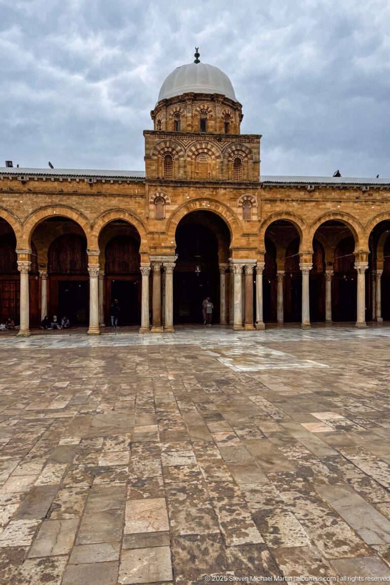 Wide paved courtyard facing the Zitouna Grand Mosque prayer-hall arcade and central domed bay beneath an overcast sky.