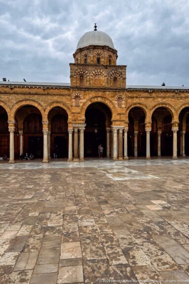 Wide paved courtyard facing the Zitouna Grand Mosque prayer-hall arcade and central domed bay beneath an overcast sky.