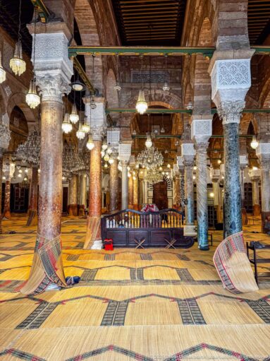 Interior of the Zitouna Grand Mosque with rows of columns and arches, hanging chandeliers, and woven prayer-floor coverings.