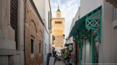 Zitouna Grand Mosque minaret above a narrow market lane with shops, pedestrians, and a green timber canopy.