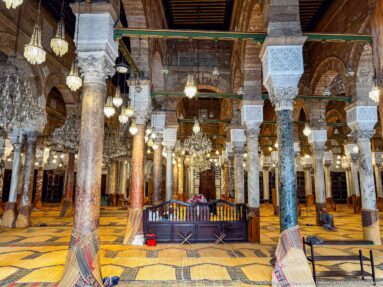 Centered view through dense rows of columns and arches inside the mosque prayer hall, with chandeliers and woven flooring.