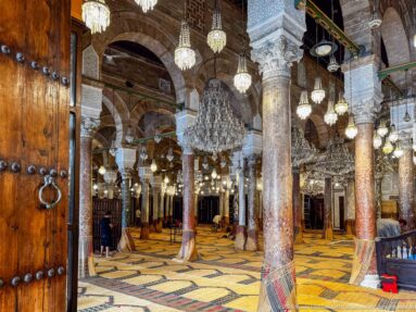 Studded wooden door with ring pull framing rows of columns, arches, chandeliers, and woven floor coverings inside the prayer hall.
