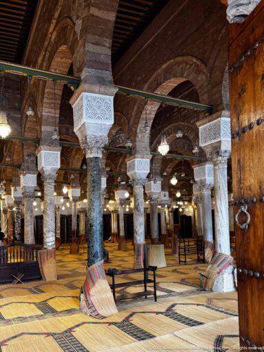 Oblique view of the Zitouna Grand Mosque prayer hall with stone columns, arches, white capitals, woven flooring, and an open wooden door.