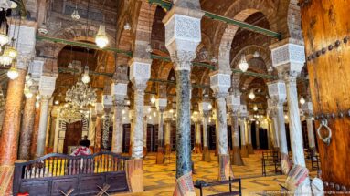 Zitouna Grand Mosque prayer hall with varied stone columns, carved capitals, striped arches, chandeliers, and a studded wooden door.