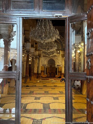 Zitouna Grand Mosque prayer hall seen through glazed doors, with a large chandelier, columns, arches, and woven floor coverings.