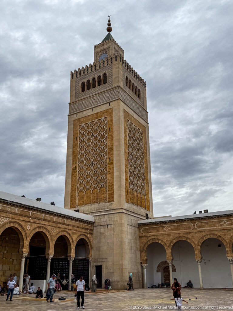 Square Zitouna Grand Mosque minaret rising above courtyard arcades, with geometric lattice panels, crenellations, and visitors below.