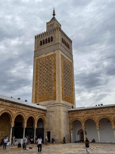 Square Zitouna Grand Mosque minaret rising above courtyard arcades, with geometric lattice panels, crenellations, and visitors below.