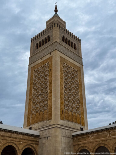 Low-angle courtyard view of the square minaret with large geometric lattice panels, upper arched openings, crenellations, and finial.
