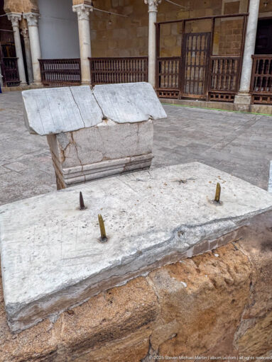 Side view of a pale stone slab with metal pegs beside a weathered carved block with a sloping cap.