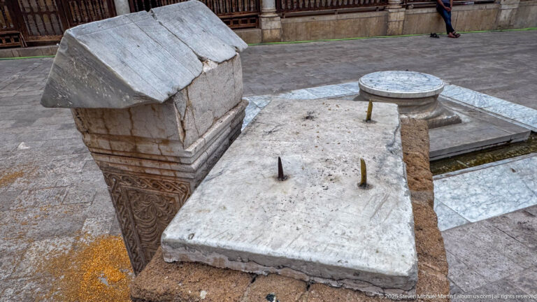 Rectangular stone slab with metal pegs beside a carved block and a low circular stone element in the mosque courtyard.