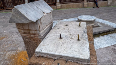 Rectangular stone slab with metal pegs beside a carved block and a low circular stone element in the mosque courtyard.