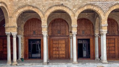 Three mosque arcade bays with carved capitals, pale columns, and large wooden screens and doors.