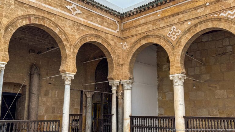 Corner of the Zitouna Grand Mosque arcade with intersecting arches, pale columns, carved capitals, and a wooden balustrade.