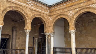 Corner of the Zitouna Grand Mosque arcade with intersecting arches, pale columns, carved capitals, and a wooden balustrade.