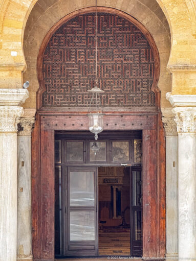 Geometric wooden lattice screen above a dark doorway, framed by pale stone and carved capitals.