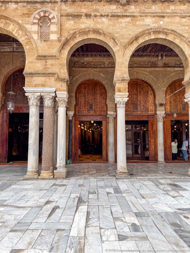 Two large courtyard arches with pale columns and open wooden doorways leading into the illuminated prayer hall.