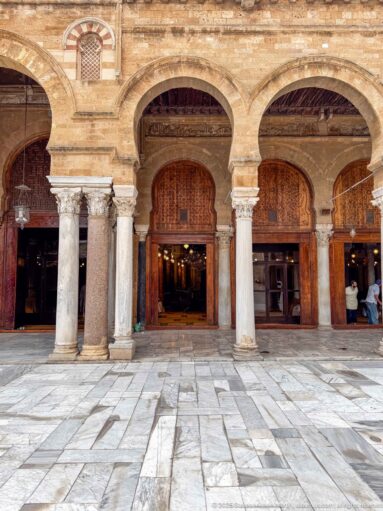 Two large courtyard arches with pale columns and open wooden doorways leading into the illuminated prayer hall.
