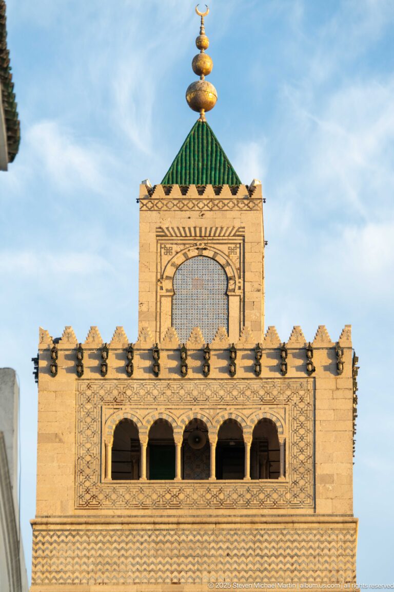 Close view of the Zitouna Grand Mosque minaret crown with geometric lattice, arched openings, green roof, and metal finial.