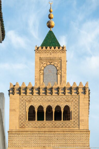 Close view of the Zitouna Grand Mosque minaret crown with geometric lattice, arched openings, green roof, and metal finial.