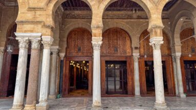 Cluster of varied stone columns beneath courtyard arches, with wooden screens and a lit prayer-hall doorway behind.