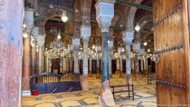 Studded wooden door framing rows of mosque columns, carved capitals, chandeliers, and woven prayer-floor coverings.