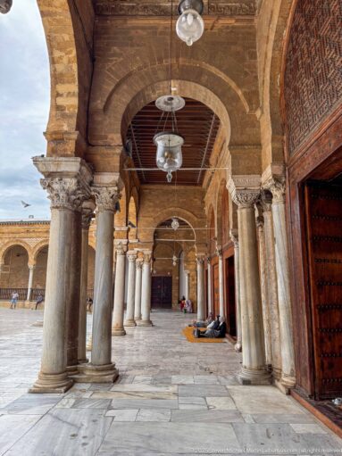 Long perspective beneath a courtyard arcade with stone columns, hanging lamps, wooden doors, and the open court at left.