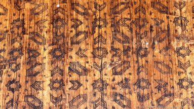 Close-up of warm wood covered with repeated incised stars, chevrons, lozenges, and angular geometric motifs.