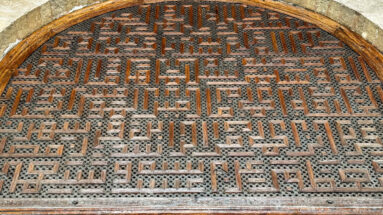 Arched wooden lattice panel filled with dense rectilinear Arabic calligraphic forms over a perforated backing.