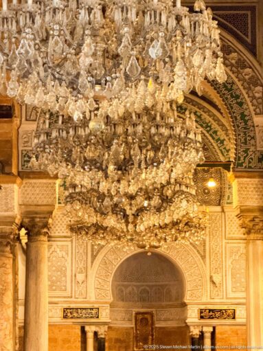 Crystal chandeliers hanging before the ornamented mihrab wall, with a recessed central niche and flanking columns.