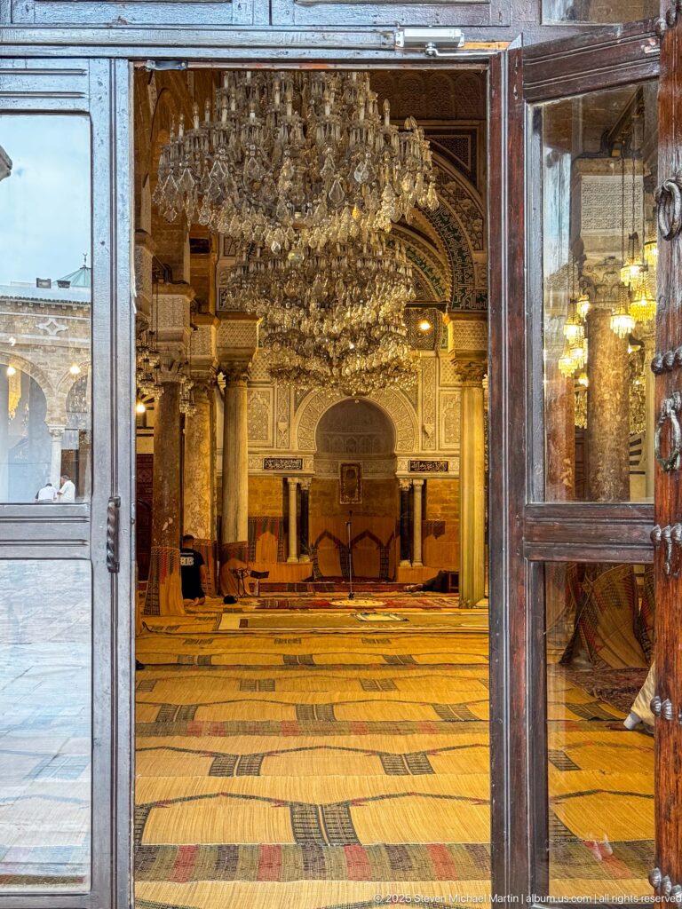 Open doorway framing the mosque prayer hall, with woven floor coverings, columns, chandeliers, and the mihrab in the distance.