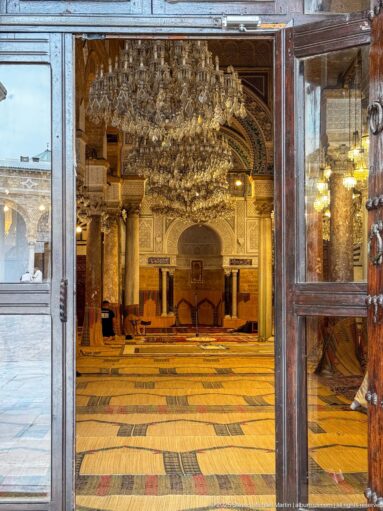 Open doorway framing the mosque prayer hall, with woven floor coverings, columns, chandeliers, and the mihrab in the distance.