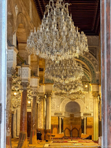 Frontal prayer-hall view with tiered crystal chandeliers aligned above the ornate mihrab and flanking columns.