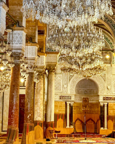 Closer view of the ornate mihrab wall beneath crystal chandeliers, with polished columns and patterned arches.