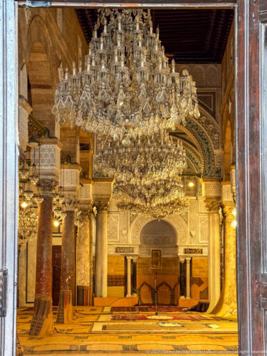 Dark wooden doorway framing a symmetrical view of chandeliers, columns, patterned flooring, and the mihrab beyond.
