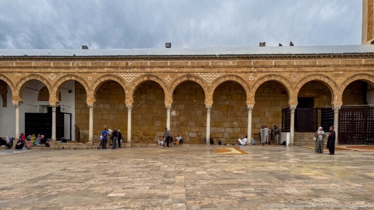 People sitting and walking beneath a long stone arcade along the Zitouna Grand Mosque courtyard.