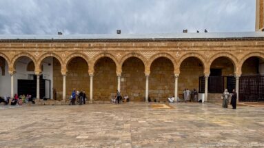 People sitting and walking beneath a long stone arcade along the Zitouna Grand Mosque courtyard.