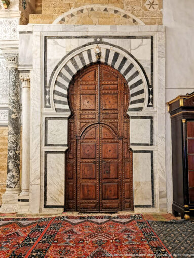 Carved wooden double doors beneath a black-and-white striped horseshoe arch, with marble framing and red patterned carpets.