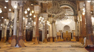 Long central aisle through the mosque prayer hall, with polished columns, chandeliers, lamps, woven flooring, and the mihrab beyond.