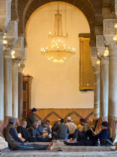 Group of men seated on floor coverings beneath a chandelier between stone columns inside the Zitouna Grand Mosque.
