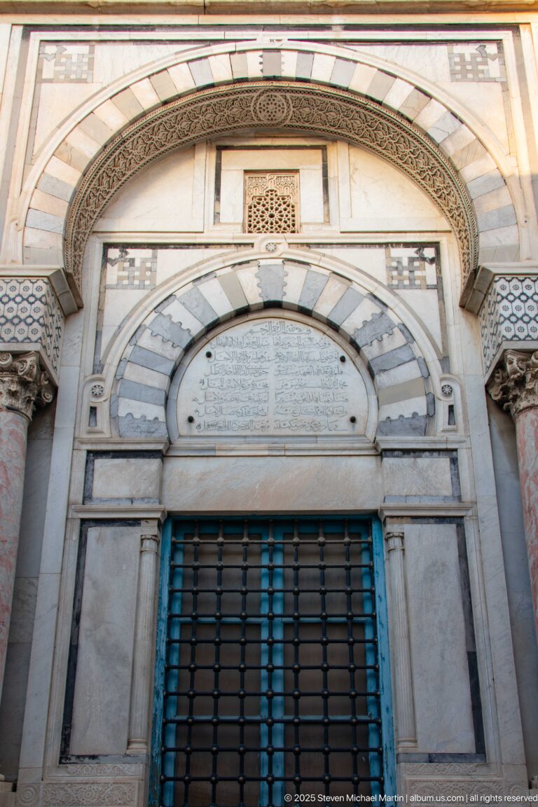 Arched stone portal with striped voussoirs, Arabic inscription panel, carved borders, small columns, and metal grille.