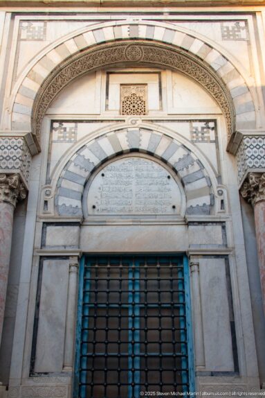 Arched stone portal with striped voussoirs, Arabic inscription panel, carved borders, small columns, and metal grille.