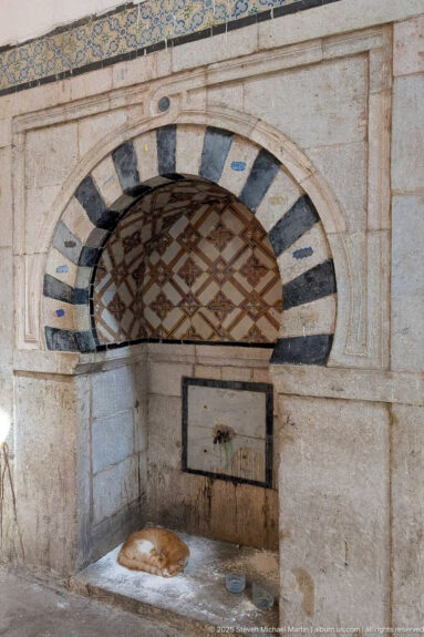 Striped arched wall niche with geometric grille, small water outlet, mineral staining, and a low stone basin.
