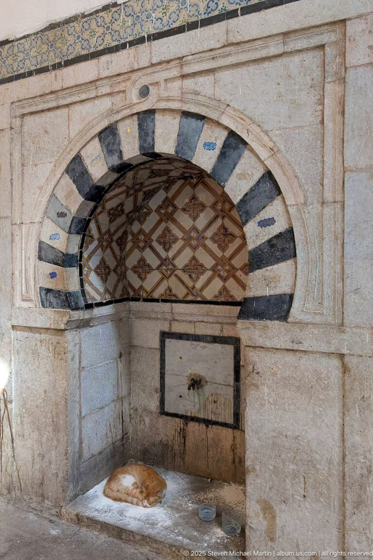 Striped arched wall niche with geometric grille, small water outlet, mineral staining, and a low stone basin.