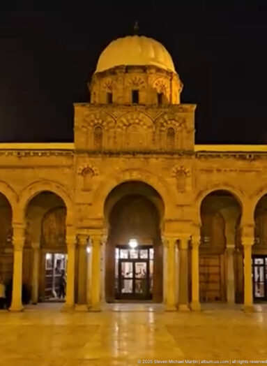 Illuminated Zitouna Grand Mosque courtyard façade at night, with horseshoe arches, columns, and a domed central bay.