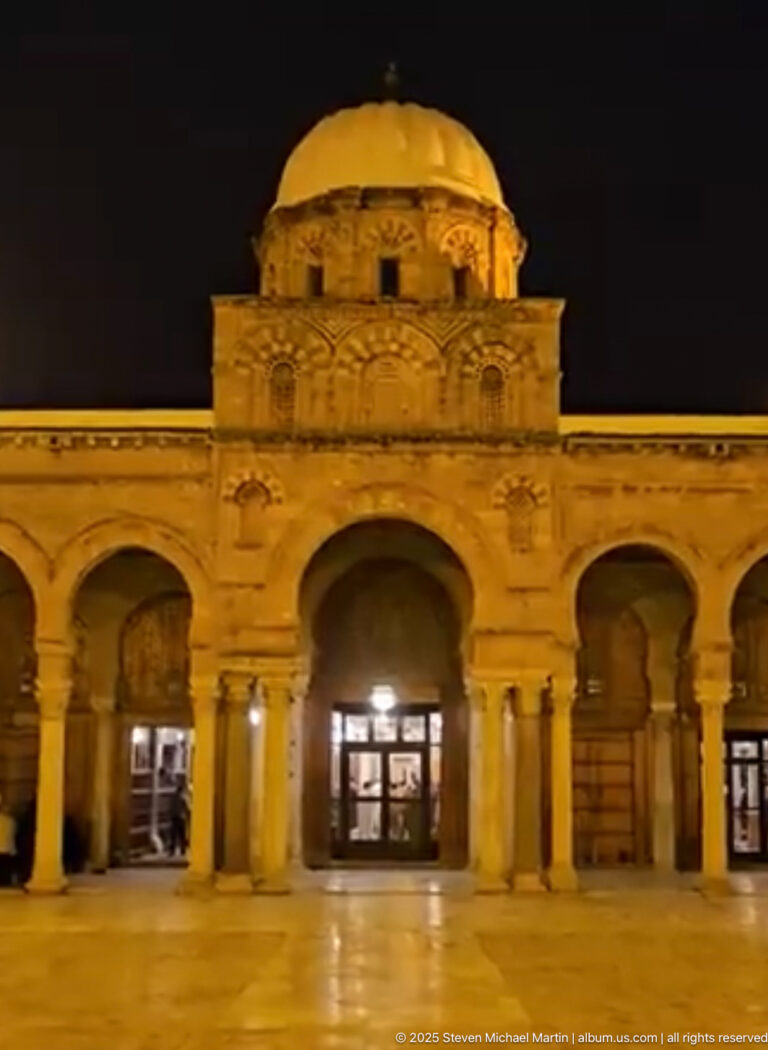 Illuminated Zitouna Grand Mosque courtyard façade at night, with horseshoe arches, columns, and a domed central bay.
